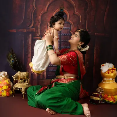A joyful mother lifts her baby, dressed as little Krishna, into the air. This playful interaction against a traditional backdrop captures the heart of the Yashoda-Krishna story.