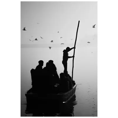 A black and white silhouette of a boatman steering his vessel through the foggy waters of the Yamuna. The image has a timeless, serene quality, capturing the quiet dedication of his work.