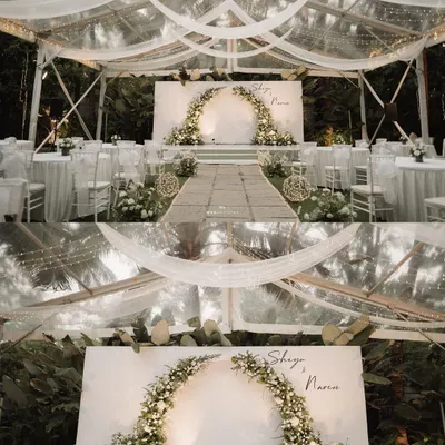 A serene and intimate wedding reception setup inside a transparent tent. The decor is all-white, featuring a beautiful floral arch, clean tablescapes, and elegant draping that creates a dreamy, romantic atmosphere.