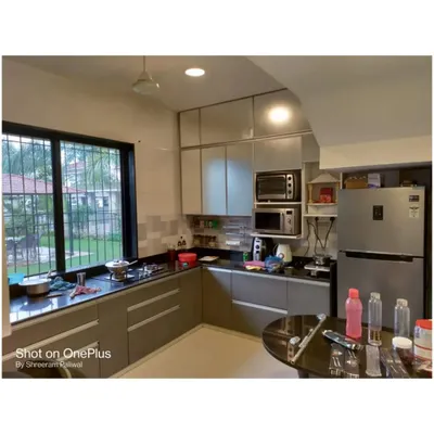This L-shaped kitchen features laminate cabinets with sleek profile handles and a dark granite countertop. The layout is designed for practicality, with the refrigerator and microwave easily accessible.