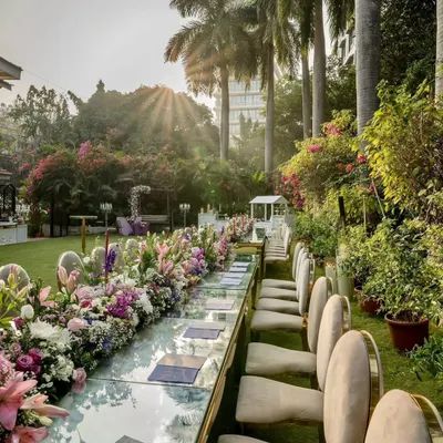A wide view of an outdoor corporate gala setup, featuring a long dining table with a lush floral runner. The sunlit garden setting at this Mumbai venue provides a beautiful, natural backdrop for a memorable event.