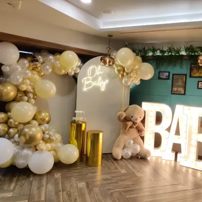 A full view of a classic "Oh Baby" shower setup. This popular theme includes a white and gold balloon garland, an arched backdrop with a neon sign, a large teddy bear prop, and light-up "BABY" letters.