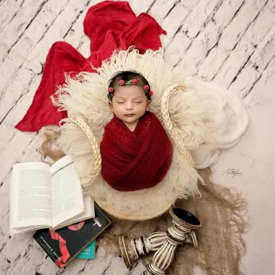 A beautifully composed overhead shot of a newborn wrapped in a rich red knit, sleeping in a basket surrounded by books and vintage decor.