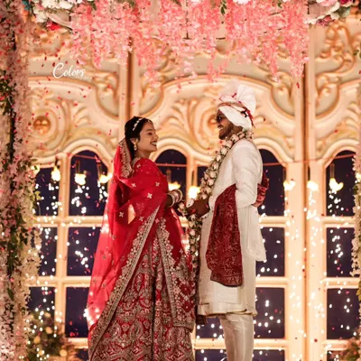 A breathtaking Varmala moment on an elevated stage, set against a stunning floral backdrop and a shower of petals. This is a true fairytale shot.