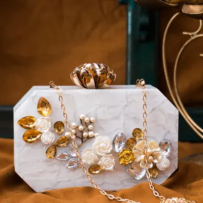 This elegant white marble acrylic clutch is adorned with golden and white floral details, pearls, and crystals. It comes with a delicate gold chain for easy carrying.