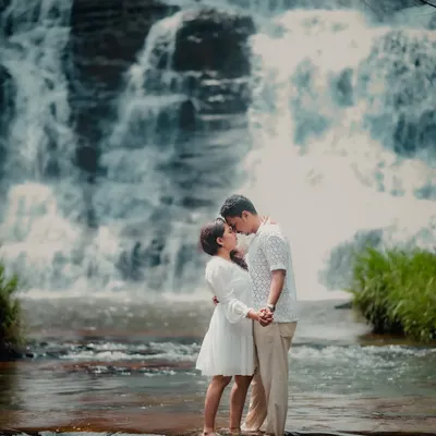 A truly epic and romantic pre-wedding moment. The couple shares a kiss while standing on a rock in the middle of a river, with a magnificent waterfall cascading behind them.