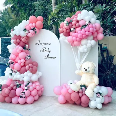 An elegant and sophisticated baby shower theme. This beautiful outdoor setup by the pool features a teddy bear holding a bouquet of pink and white balloons, with a matching organic balloon arch against a clean white backdrop.
