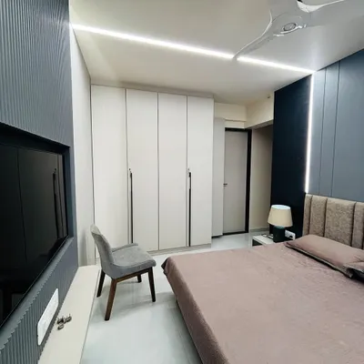 A wide view of a modern bedroom featuring a floor-to-ceiling white wardrobe and a contrasting dark grey fluted panel behind the TV. The linear ceiling lights provide clean, even illumination.