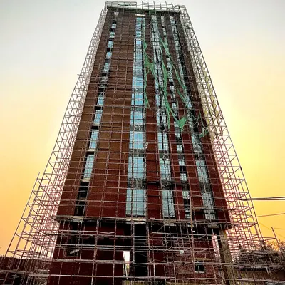 A full view of a residential tower under construction, showcasing the extensive use of exposed brick across its facade. The scaffolding reveals the scale of the project and the meticulous nature of the brickwork in progress.