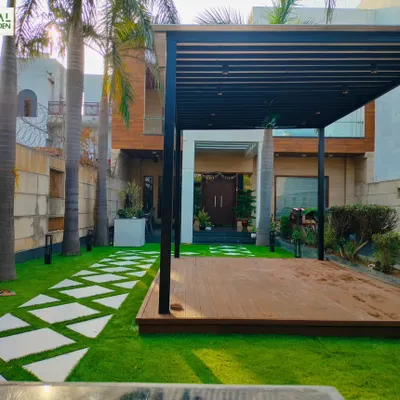 A completed front yard project in Dwarka, New Delhi, featuring a large wooden deck under a black pergola, surrounded by a manicured lawn and geometric pathway.