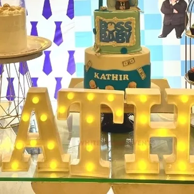 A close-up of custom light-up marquee letters spelling a name, placed on a glass table in front of a "Boss Baby" themed birthday cake.