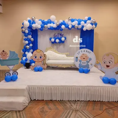 This stage decoration for a baptism ceremony features a blue and white balloon arch over drapes, with a comfortable sofa for the family. Themed cutouts and a circular sign add a personalized touch to the setup.