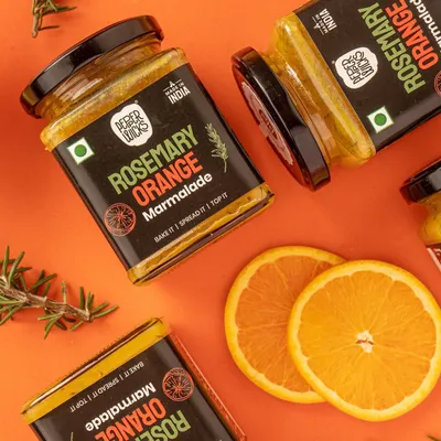 A dynamic flatlay for Pepper Wicks' Rosemary Orange Marmalade. The scattered jars, fresh orange slices, and rosemary sprigs clearly communicate the product's ingredients and flavor profile in a visually exciting way.