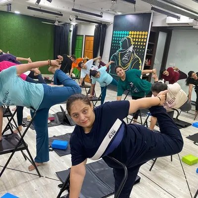 A full class of smiling faces enjoying a supported balancing pose in Chair Yoga. This practice is perfect for building stability and confidence at any age.