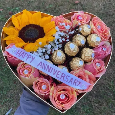 A heart-shaped anniversary box celebrating love with a bright sunflower, delicate pink roses, and a cluster of Ferrero Rocher chocolates. The personalized "Happy Anniversary" ribbon completes this thoughtful gift.