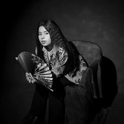 A moody black and white portrait. The shadows and the model's pensive expression, along with the fan prop, create a cinematic and artistic image.