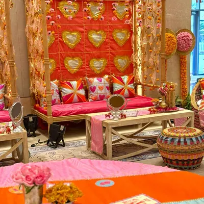 A festive seating arrangement for a Garba celebration, featuring a wooden four-poster canopy. The backdrop is a vibrant pink quilt with golden heart motifs, complemented by colorful custom cushions and traditional decor elements.