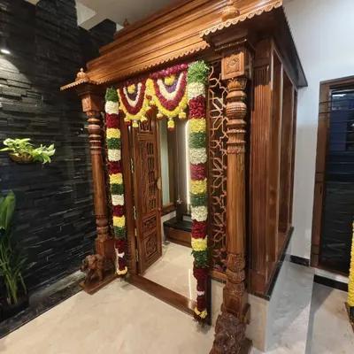A stunning traditional wooden pooja room entrance, beautifully decorated with thick garlands of fresh flowers for a housewarming ceremony.
