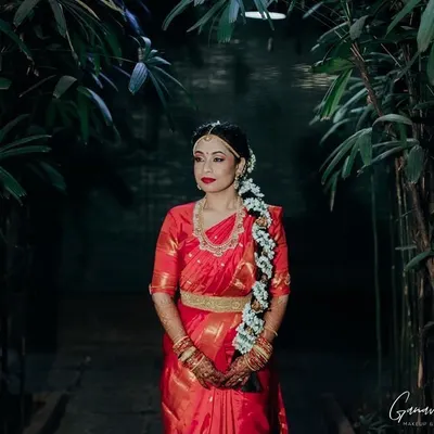 This is a classic South Indian bridal portrait. The makeup features a bold red lip and defined eyes, perfectly complementing her traditional red saree, temple jewelry, and jasmine-adorned braid for a timeless look.