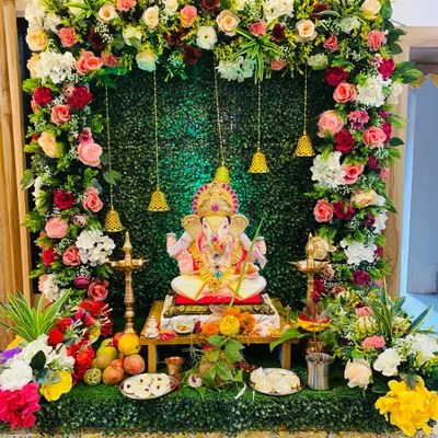 This modern Ganpati setup features a rich arch of fresh roses against a verdant green wall. The hanging brass bells and traditional lamps blend perfectly with the contemporary floral design, creating a truly divine atmosphere.