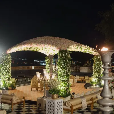 A stunning floral dome mandap set against the Udaipur city palace skyline at night. The structure is supported by pillars wrapped in white flowers, creating an open and airy feel for the wedding ceremony.