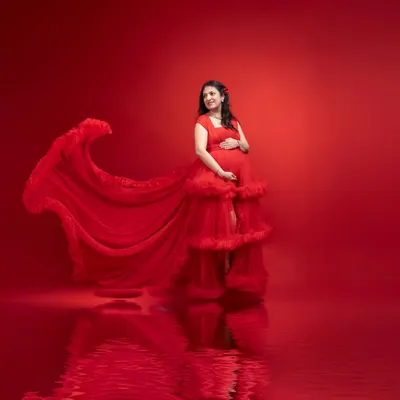 For a truly showstopping memory, our studio maternity sessions offer a high-fashion experience. This portrait features a stunning red tulle gown with a dramatic train, set against a matching backdrop with a beautiful reflection. The professional lighting highlights the mother's joyful expression and the beautiful silhouette of her baby bump.