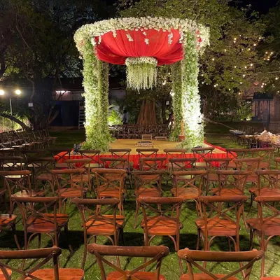A bold and beautiful outdoor mandap at night, featuring pillars of greenery, a vibrant red canopy, and a chandelier of white flowers, surrounded by rustic wooden chairs.
