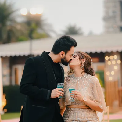 A candid moment from a cocktail party or reception. The couple shares a kiss while holding drinks, capturing the relaxed and celebratory vibe.