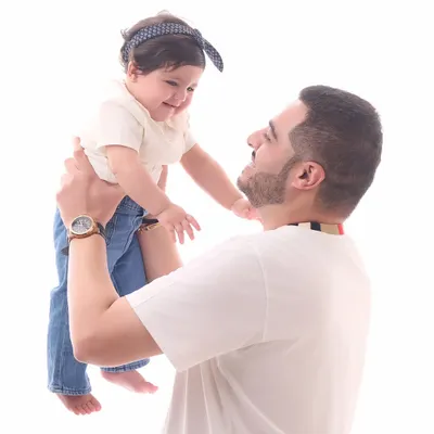 A classic father-daughter portrait against a clean white background. The father lifts his baby girl, capturing a moment of pure joy and connection in our bright and timeless style.
