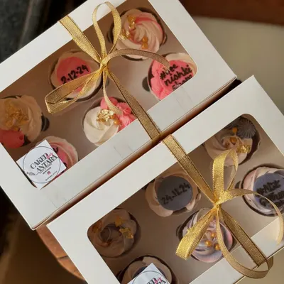 Two beautiful boxes of cupcakes ready for a bridal shower. The designs feature custom dates and names, with delicate pink and purple frosting and gold ribbon.