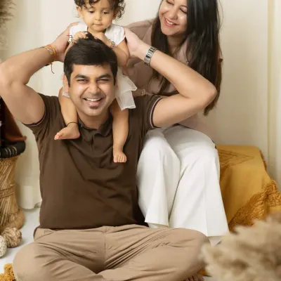 A playful studio family portrait where a father gives his daughter a shoulder ride, with mom looking on with a huge smile.