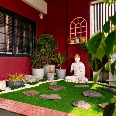 A balcony in Faridabad transformed into a tranquil Zen garden. The space features a serene Buddha statue, artificial grass with stone pavers, and a variety of potted plants against a bold red wall.