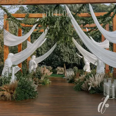 The grand entrance to the 'Ivory Garden' reception, featuring a wooden pergola draped with flowing white fabric and greenery. Classical statues line the wooden walkway, creating a dramatic and romantic welcome for guests.