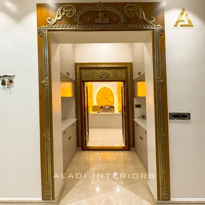 A grand entrance to a puja room, framed by an ornate, traditionally carved wooden archway. The inner doors feature intricate brass work, leading to a sanctum with a backlit onyx-style panel.