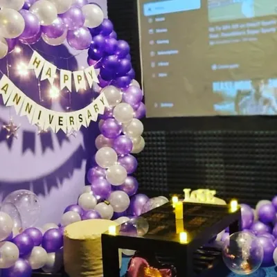 A beautiful anniversary setup with a purple and white balloon arch and a 'Happy Anniversary' banner. The soft lighting from the fairy lights adds a romantic touch to the room.