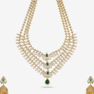 A luxurious three-layer necklace set made with American Diamonds and emerald-colored stones. The set includes matching jhumka earrings, creating a complete and opulent look for a bride or wedding guest.