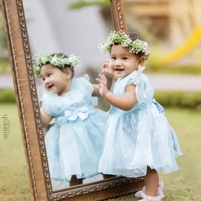 An adorable baby girl in a light blue dress and a floral crown looks at her reflection in a large, ornate mirror during an outdoor photoshoot. This creative setup makes for a magical portrait.