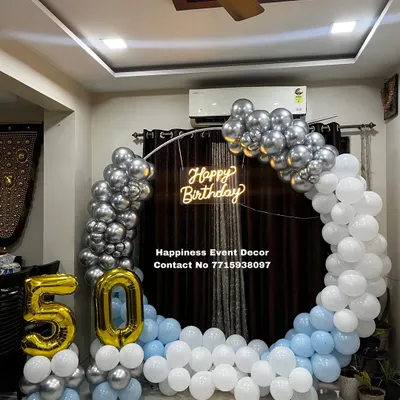 A stylish 50th birthday home decoration. This setup features a balloon half-arch in silver, white, and blue, a 'Happy Birthday' neon sign, and large foil balloons for the number '50'.
