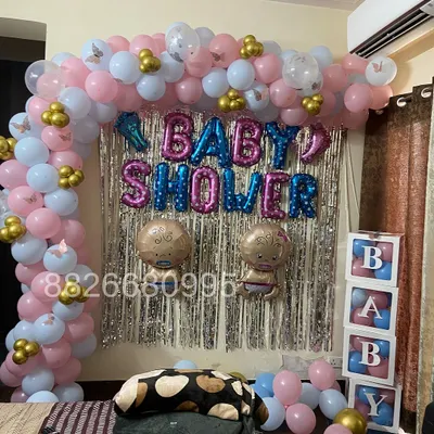 A complete baby shower decoration for a home celebration. The setup includes a balloon arch in pink, light blue, and gold, a silver fringe backdrop, and 'BABY' blocks on the side.