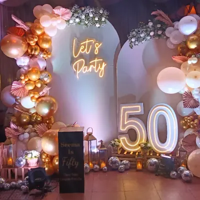 A fabulous 50th birthday celebration! This setup features a "Let's Party" neon sign, large marquee numbers, and a stunning balloon arrangement in rose gold, white, and gold.