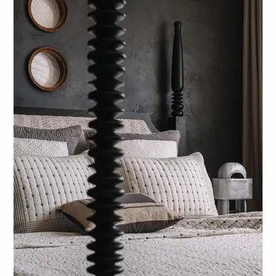 The master bedroom in our 'Aria' project uses a stark, masculine palette of black and white with bursts of mud red. A custom black burnt-wood four-poster bed creates a dramatic centerpiece, viewed here through its intricately turned post, adding a layer of sculptural detail.
