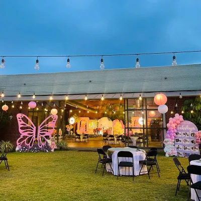 A wide shot of an enchanted garden party setup in a spacious backyard. The scene features a large pink butterfly prop, string lights, and themed seating areas for guests.
