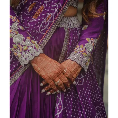 A close-up on the exquisite details of the bride's purple bandhani-style lehenga. The intricate embroidery and delicate henna on her hands tell a story of tradition and craftsmanship.