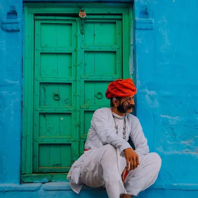 A man with a magnificent mustache and a red turban sits against a vibrant blue wall with a green door in Jodhpur. This is the quintessential color palette of Rajasthan, bold and full of character.