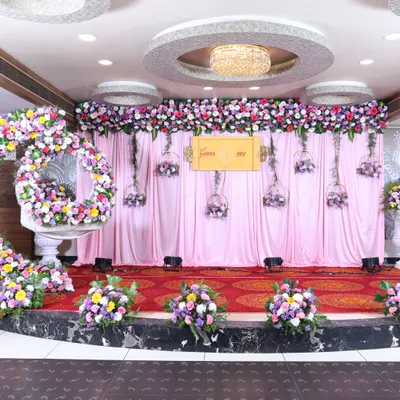 A large-scale stage decoration for a reception or engagement. The setup includes a long floral border across the top, hanging floral baskets, and a large, circular floral arrangement for photos.