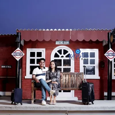 A travel-themed pre-wedding shoot using the vintage railway station set. The couple is dressed in casual attire, sitting on a bench with luggage props to complete the scene.