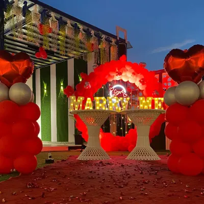 A beautiful daytime view of a rooftop proposal decoration. The setup includes bold red balloon pillars with heart toppers, a red carpet aisle, and large illuminated "MARRY ME" letters, all ready for that magical moment.