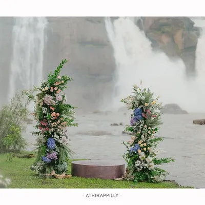 The stage is set for a vow renewal ceremony at the majestic Athirapilly waterfalls. The simple, elegant decor with floral pillars stands in beautiful contrast to the raw power of nature, creating a truly unique wedding venue.