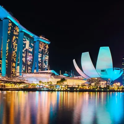 The iconic Marina Bay Sands and ArtScience Museum in Singapore are brilliantly lit in blue and white, their reflections shimmering on the water at night.