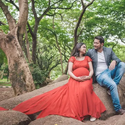 A serene moment between an expecting couple, sitting on a large rock formation in a park, surrounded by greenery. The flowing red gown adds a beautiful pop of color.